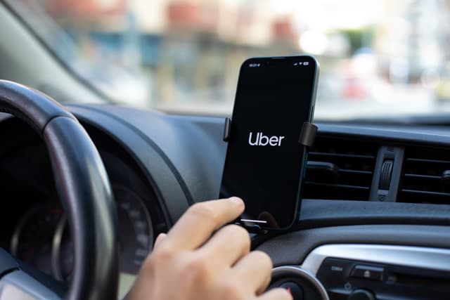 Close-up of a driver’s hand tapping a smartphone mounted on a car dashboard with the Uber app open on the screen.