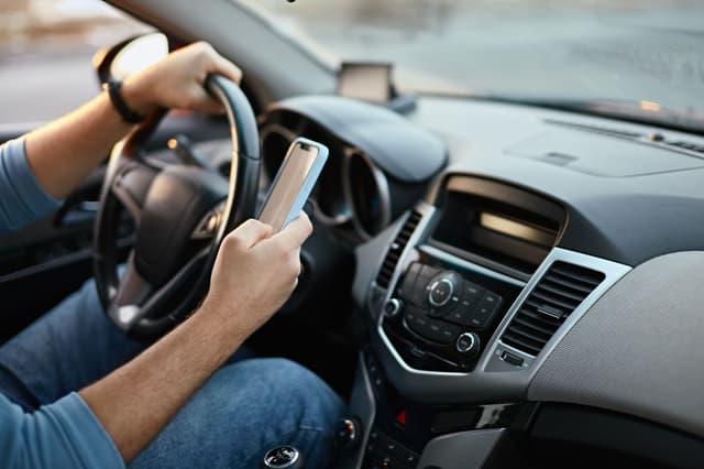 A driver holding a smartphone while operating a car, with one hand on the steering wheel and the other on the phone, illustrating the dangers of distracted driving.