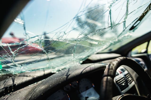 Close-up view from inside a car showing a shattered windshield and damaged steering wheel after a crash, with sunlight reflecting off the broken glass.