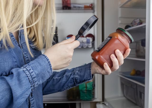 A woman holding a jar of food in front of an open refrigerator while using a magnifying glass to read the product label.